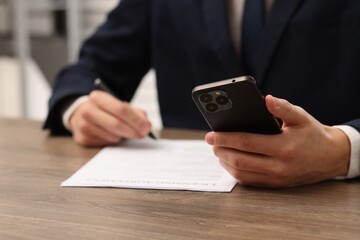 Man with smartphone signing licensing agreement document at wooden table indoors, closeup