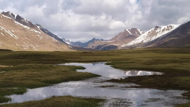 Stunning footage of a lake at Arabel Plateau with dry grass, greenery, snowy mountains, and cloud reflections.