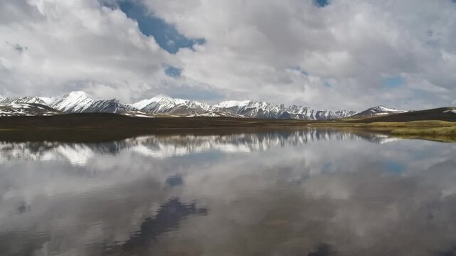 Drone footage of a lake reflecting mountains and clouds with shifting sunlight, filmed at Arabel Plateau, Kyrgyzstan.
