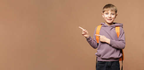 Cute schoolboy with backpack pointing at something on dark beige background, space for text. Banner