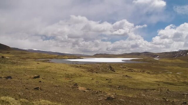 A time-lapse 4K shot of Arabel Plateau in Kyrgyzstan showing a frozen lake surrounded by lush meadows, with clouds moving rapidly across the sky, creating a dynamic atmosphere