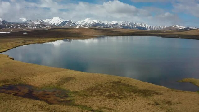 Footage of a picturesque lake in Arabel Plateau, Kyrgyzstan, surrounded by lush greenery and snowy mountain peaks, with dynamic weather shifting between cloudy and sunny skies