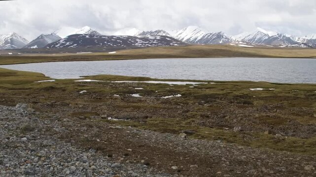 Drone footage showcasing the stunning Arabel Plateau in Kyrgyzstan with a lake surrounded by vast grass plains and snowy mountain peaks in the background. Perfect for nature and landscape enthusiasts