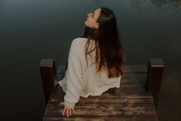 A woman sits on a wooden dock by a calm body of water, looking over her shoulder. She wears a beige sweater, black scarf, and polka-dot skirt, enjoying the serene outdoor atmosphere.