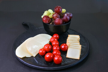 Assorted snacks with fresh grapes, cheese slices, square crackers, and cherry tomatoes, beautifully arranged on a black plate. Perfect for appetizers or a light meal.