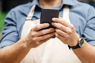 Sales plants, decor, gifts and startup. Busy millennial guy in apron and protective mask typing message or surfing on internet in social networks on smartphone near flower shop, close up, cropped