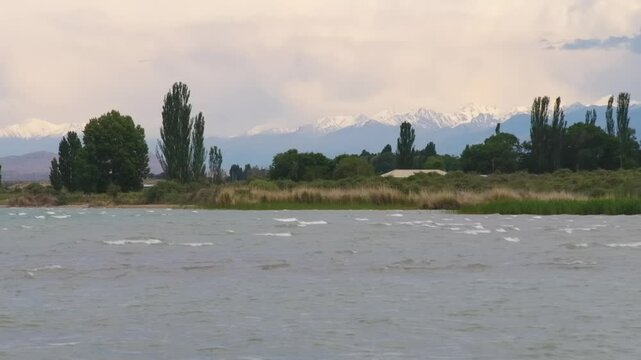 Footage showcasing the beautiful Assyk Kul Lake with trees lining the shore and snowy mountain peaks in the background, perfect for nature lovers and adventure seekers in Kyrgyzstan.
