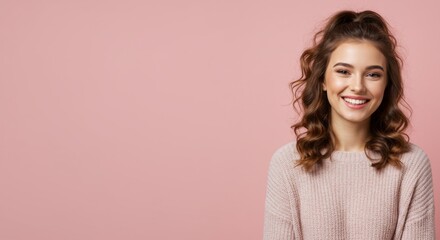 Fototapeta premium Smiling young woman with curly hair wearing a cozy sweater against a soft pink background, radiating happiness and warmth