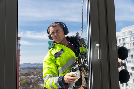 Professional Window Cleaner Working at Height in Bright Safety Gear: Focused Worker Safely Cleaning Glass at High Rise Building, Scenic Cityscape Visible Behind Him