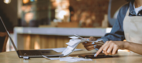 Payment of bills, bankruptcy and modern cafe during covid-19 quarantine. Millennial african american woman bartender in apron sits at table and works with laptop in interior of cafeteria, cropped