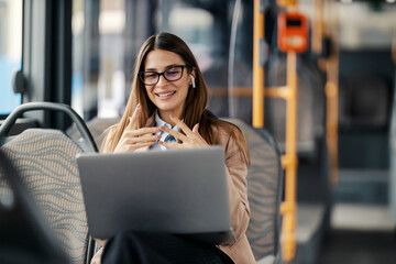 Portrait of young happy fashionable lady sitting in city bus with laptop and having video call.