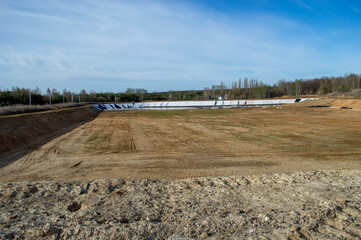 A vast construction site shows an empty sports arena with uneven earth and a clear blue sky in a...