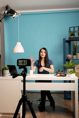 Beauty influencer sits at desk in a studio with a blue backdrop, engaging with her online audience during livestream. Perfect for themes of vlogging, content creation, and influencer marketing.
