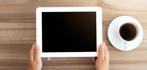 Overhead shot of unrecognizable lady using white digital tablet with isolated black screen, coffee cup on wooden table