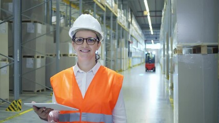 A woman in a hard hat and orange vest stands in a warehouse, holding a tablet. The shot emphasizes her role in logistics and safety.