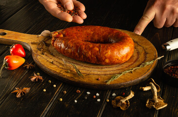 A person adds spices to a large, smoked sausage resting on a wooden cutting board. Surrounding...