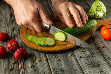 A person skillfully cuts a fresh cucumber into thin slices on a wooden cutting board. Nearby, bright red radishes and a tomato lay, adding color to the rustic kitchen setting