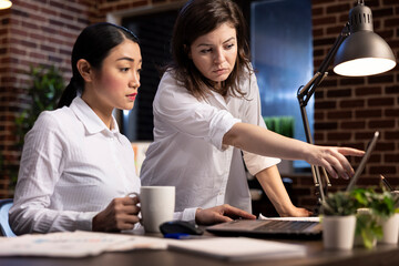 Productive night meeting between diverse businesswomen as they work together, reviewing company plans and making decisions in office. Caucasian manager pointing at laptop as she talks to her employee.