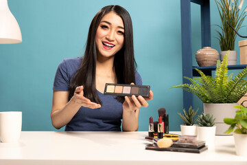 Smiling beauty influencer with eyeshadow palette, recording a cosmetics tutorial, presenting makeup...