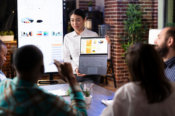 Asian financial advisor confidently leads a workplace discussion, presenting financial data on laptop. The modern office setting, digital screen and engaged team reflects dynamic business environment.