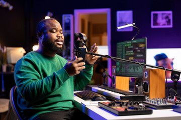 Talented young artist performing an acoustic version of his new song, reading the lyrics on his smartphone. Professional musician producing a new track, recording audio files on microphone.