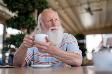 Old man with a long white beard sitting in a cozy café, holding a cup of coffee while gazing into...