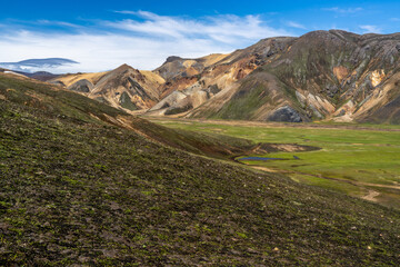 A panorama scene of colorful volcanic landscape with a moss-covered slope leading to a green valley and brown mountains along the Laugavegur trail, Iceland