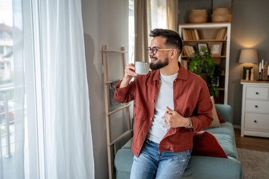 Young man drinking coffee and looking out the window in his apartment