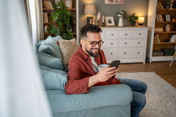 Smiling man using smartphone and drinking coffee on sofa at home