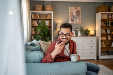 Fototapeta premium Young man recording voice message while drinking coffee on sofa at home