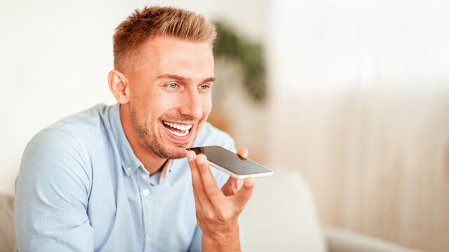 Voice Search Concept. Closeup portrait of young bearded guy using voice assistant or calling on smartphone at home, blurred background, copy space. Smiling man talking on mobile phone