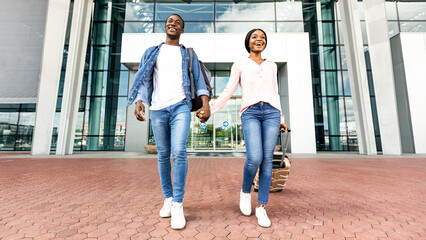 Fototapeta premium Ready For Adventures. Excited Black Couple Leaving Airport Building In New Country, Enjoying Travelling Together, Holding Hands