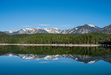 View of the Eibsee lake with a perfect reflection of the snow-capped mountains and pine forest in the calm water. The bright blue sky and natural symmetry create a feeling of calm and purity.