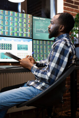 Focused stockbroker with clipboard, studying forex market trends at night, using multiple monitors...