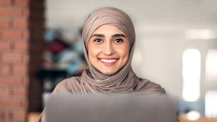 Portrait of pretty muslim girl in hijab using laptop at cafe, smiling at camera