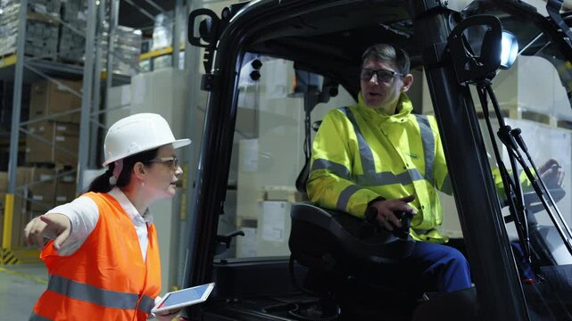 In a warehouse, a woman in an orange vest points at a man in a yellow jacket operating a forklift, indicating a discussion on logistics and storage organization.