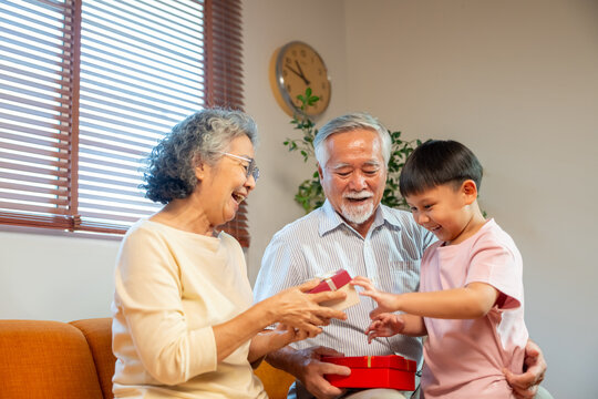 Happy two generation Asian family Grandparent and cute grandchild kid spending time together at home. Little boy giving present in red gift box to grandmother and grandfather in living room at home.