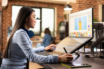 Professional female illustrator using stylus pen on graphics tablet, creating digital sketch. Asian woman working in creative agency workspace, focused on editing and designing content for production.