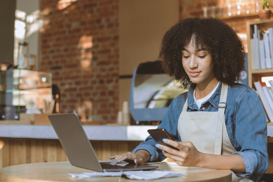 Millennial african american barista works in cafeteria with modern design loft interior maks documentation in online database. Female coffee shop cashier in apron look at phone and typing on laptop