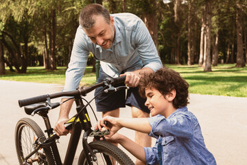 Maintenance. Close up portrait of smiling adult man and boy fixing bike using wire cutter at public park, father holding bicycle, son repairing with clippers, outdoors, selective focus