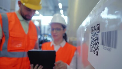 In a warehouse, two workers in orange vests and hard hats examine a package with a barcode and QR code, using a tablet for inventory management. - Powered by Adobe