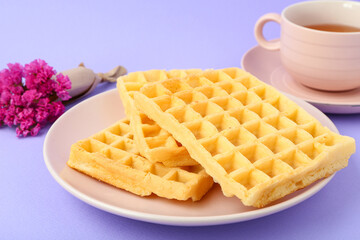 Tasty Belgian waffles in plate with cup of tea and flowers on lilac background