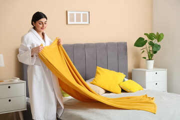 Young woman in bathrobe putting yellow blanket on bed at home