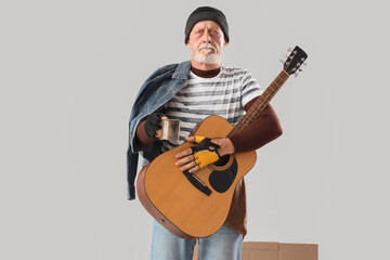 Mature homeless man with cup and guitar asking for money on light background