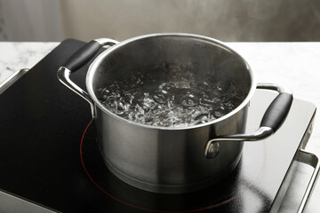 Cooking pot with boiling water and stove on light table against grey background, closeup
