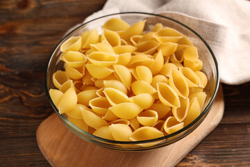 Glass bowl with raw pasta shells on wooden background