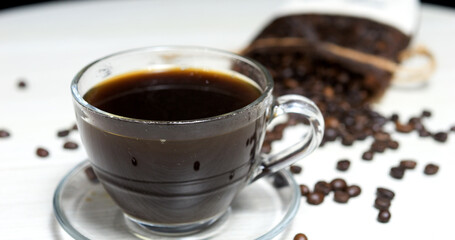 Clear glass cup filled with rich, aromatic black coffee sits on a saucer, surrounded by scattered coffee beans. The inviting scene suggests a fresh and energizing beverage