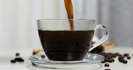Pouring rich, aromatic coffee into a clear glass cup resting on a saucer atop a white table, with...