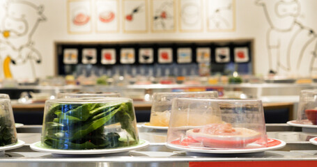 Fresh ingredients like meat, vegetables, and noodles rotate on small plates in a modern conveyor belt sushi restaurant, covered by transparent domes and ready to be picked up by customers