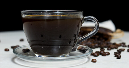 Transparent glass cup filled with invigorating black coffee resting on a glass saucer, surrounded by scattered roasted coffee beans on a white wooden table against a black backdrop